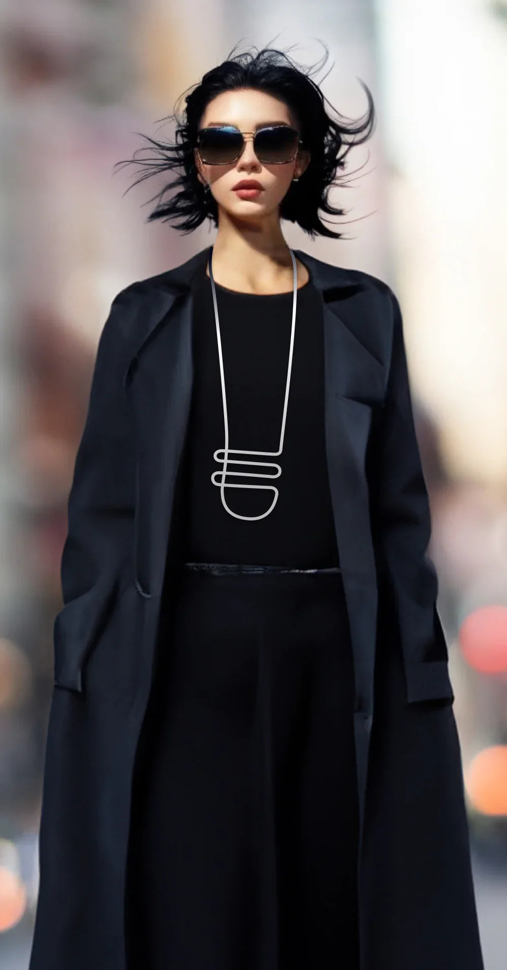 Woman wearing large sunglasses, black outfit, and long modern silver necklace in city setting
