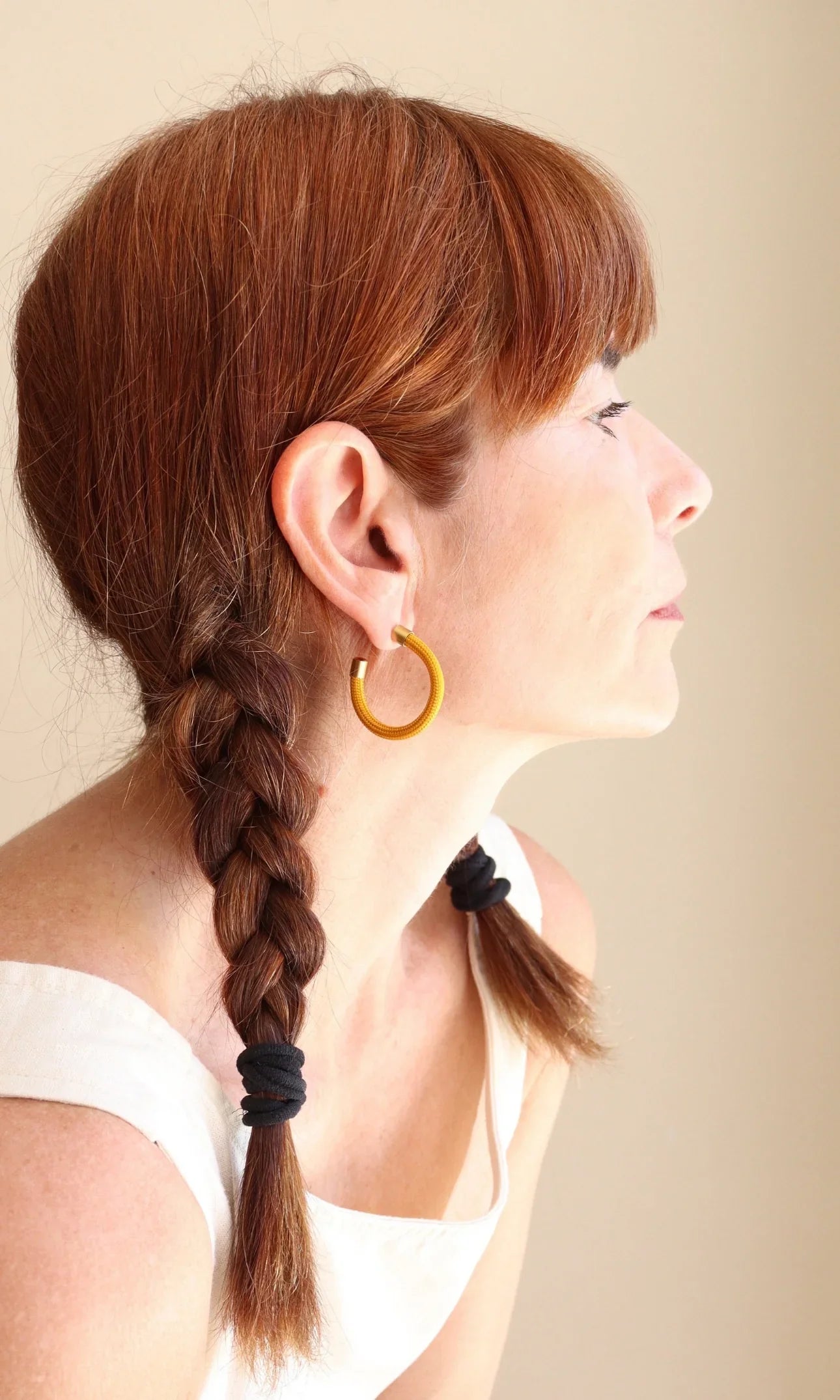 Profile of woman with red hair in braided pigtails wearing yellow hoop earrings, white top, neutral background